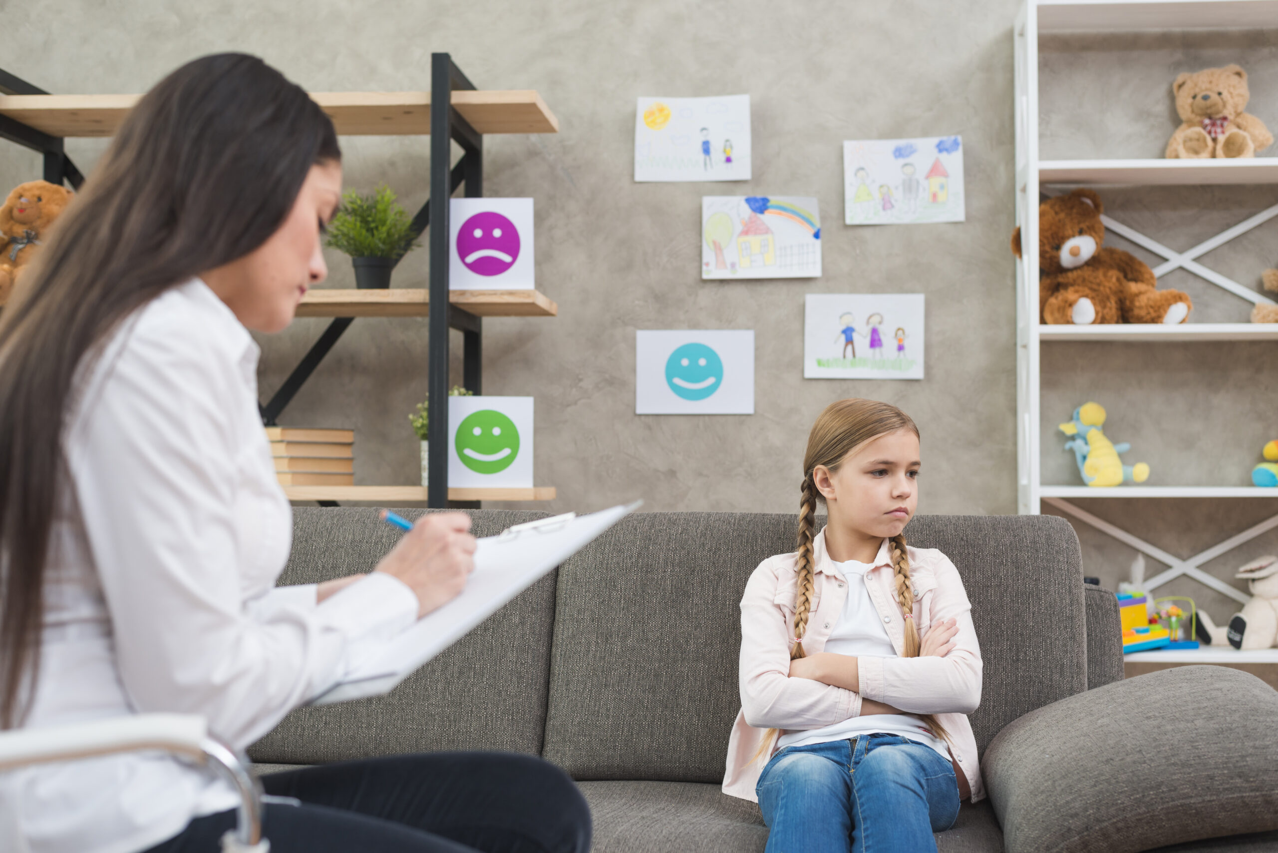 A girl sitting on a sofa with a female psychologist writing a note.