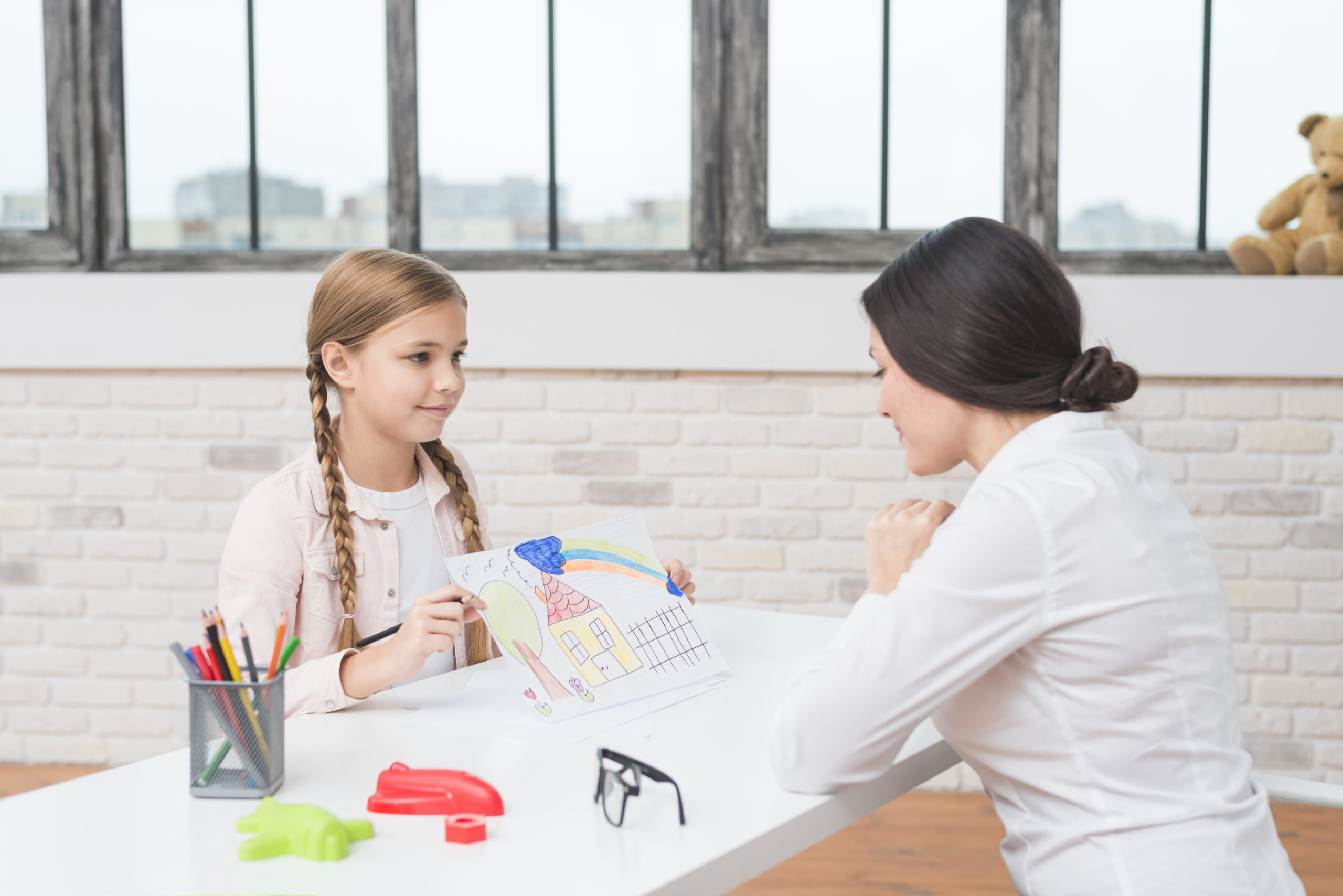 A little blonde girl showing a drawn house on paper to her female psychologist.